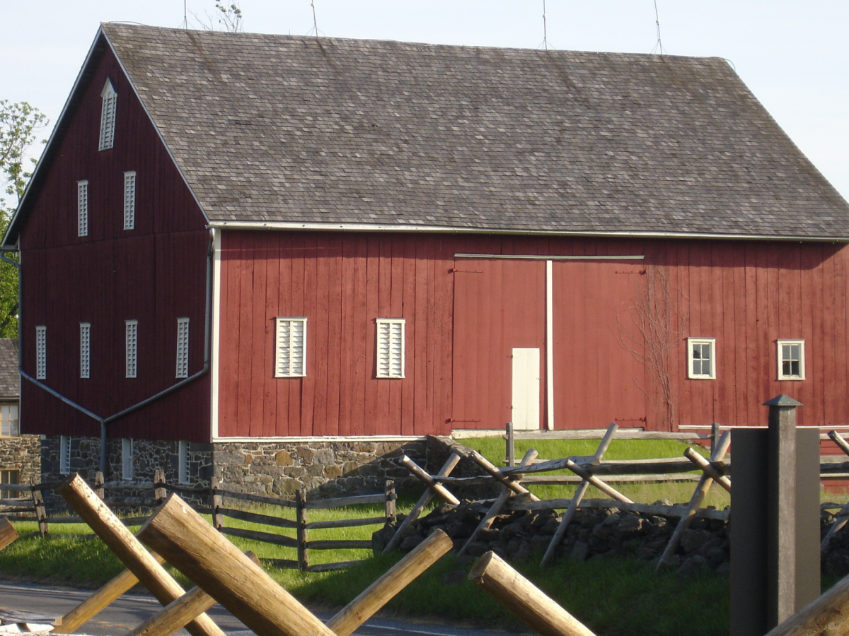 A large red barn with white-trimmed windows and a stone foundation stands behind a split-rail fence and wooden barriers, much like scenes from The Scandalous and Heroic Tale of General Daniel Sickles: Gettysburg School Trips.