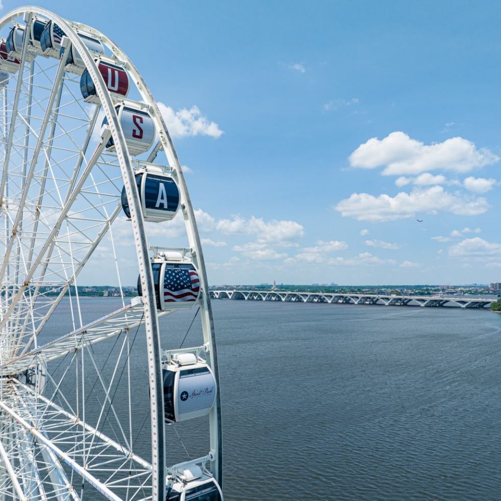 A large Ferris wheel with gondolas—one marked "USA" and another displaying the American flag—stands by the water, with a long bridge in the background under a blue sky with scattered clouds.
